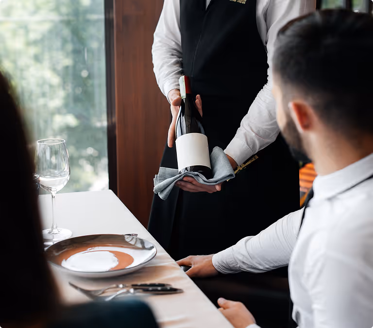Waiter holding a wine bottle wrapped in a cloth presenting it to seated diners at a restaurant table.