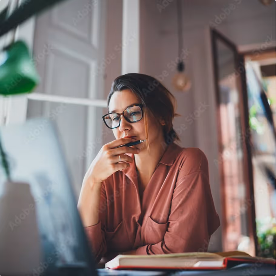 Woman in glasses sitting at a desk looking thoughtfully at a laptop with an open book nearby.
