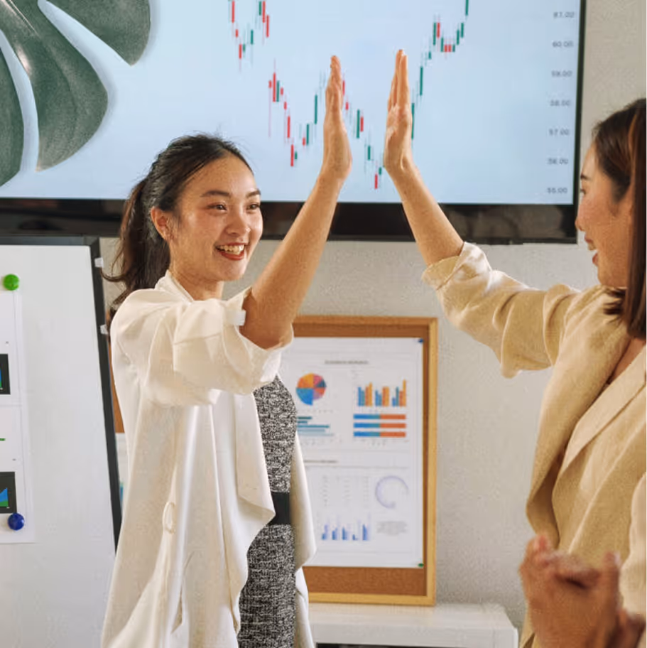 Two businesswomen high-fiving in an office with financial charts and graphs on the wall.