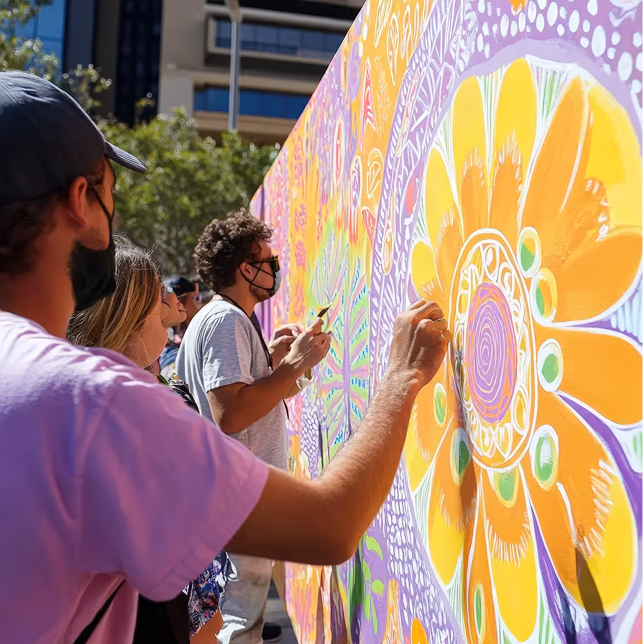 People wearing masks painting a colorful mural with large yellow flowers outdoors.