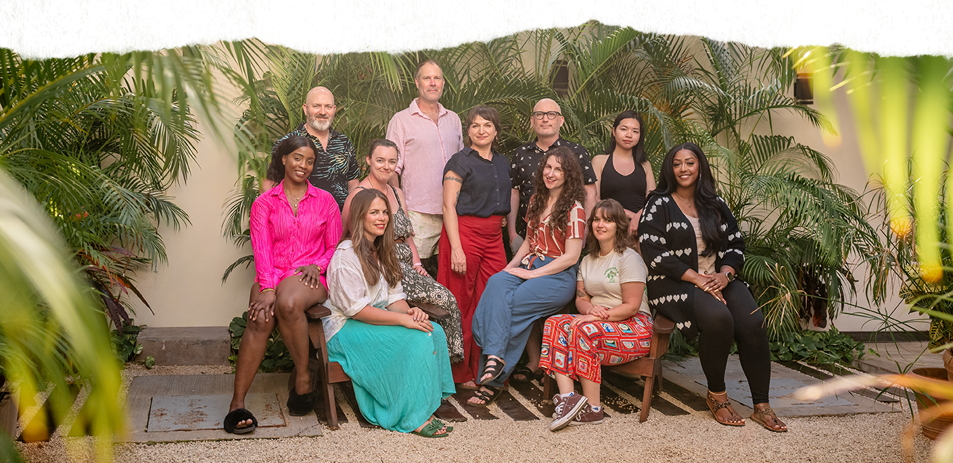 Team photo featuring eight people in a decorated indoor setting. The team members are positioned on sofas, armchairs, and the floor, smiling at the camera, with curtains, framed artwork, and decorative elements in the background.