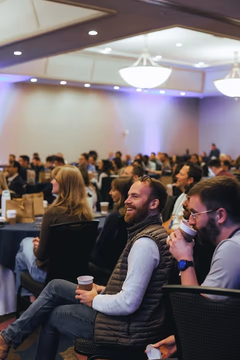 Audience seated in a conference room listening attentively, with two men in the foreground smiling and holding coffee cups.