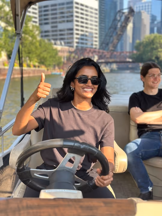 Woman wearing sunglasses smiling and giving thumbs up while steering a boat with a man sitting and smiling in the background by a river with city buildings.