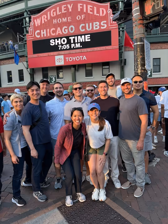 Group of smiling people posing in front of Wrigley Field sign that reads 'Home of Chicago Cubs' with a digital message 'SHO TIME 7:05 P.M.'