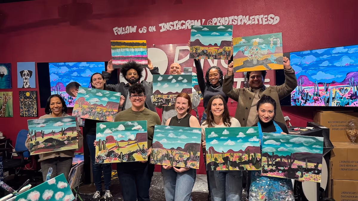 Group of people smiling and holding up landscape paintings with desert scenes and blue skies inside an art studio with colorful artwork on the walls.