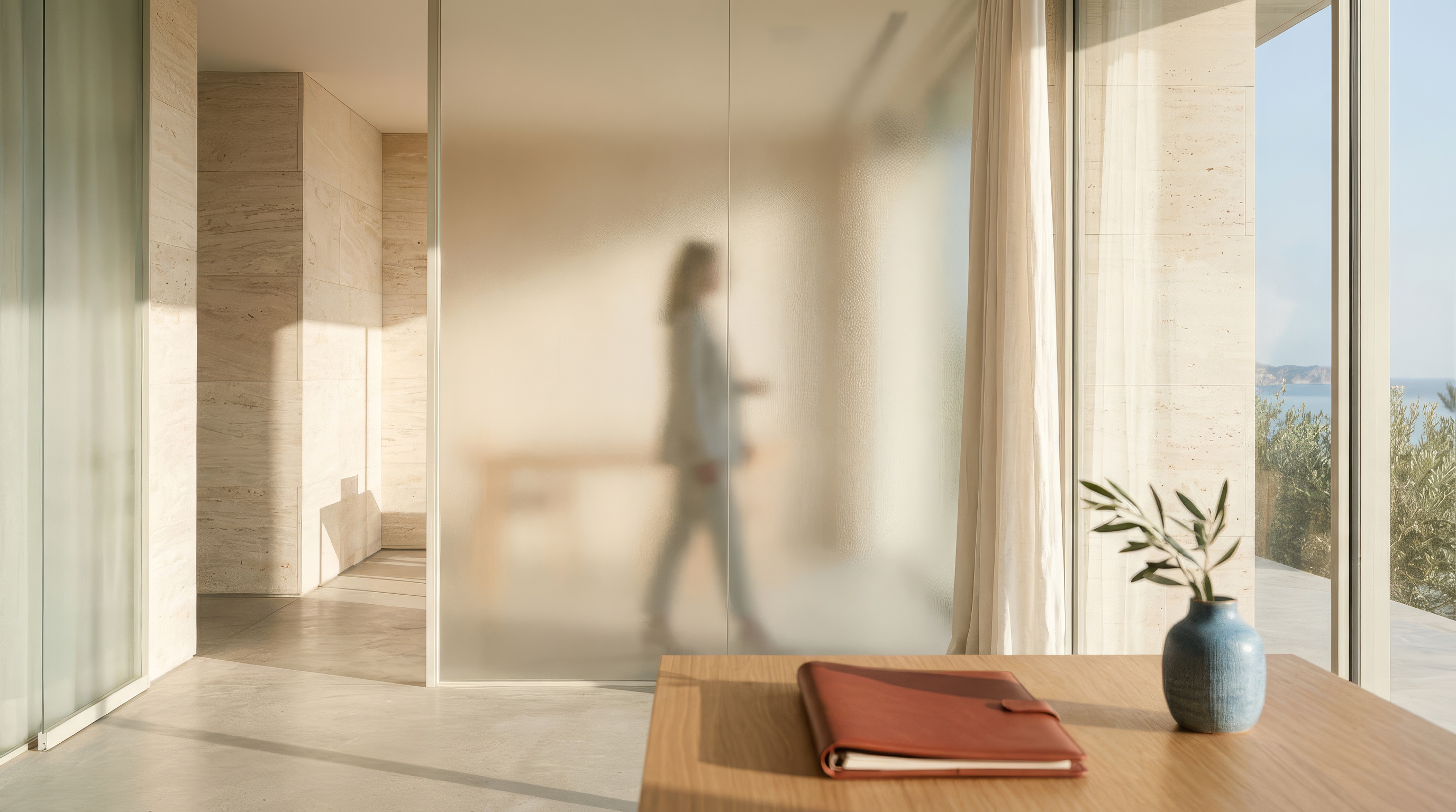 Interior moderno con luz natural, mesa de madera con cuaderno y flor en jarrón, y silueta difusa de persona tras panel de vidrio esmerilado.