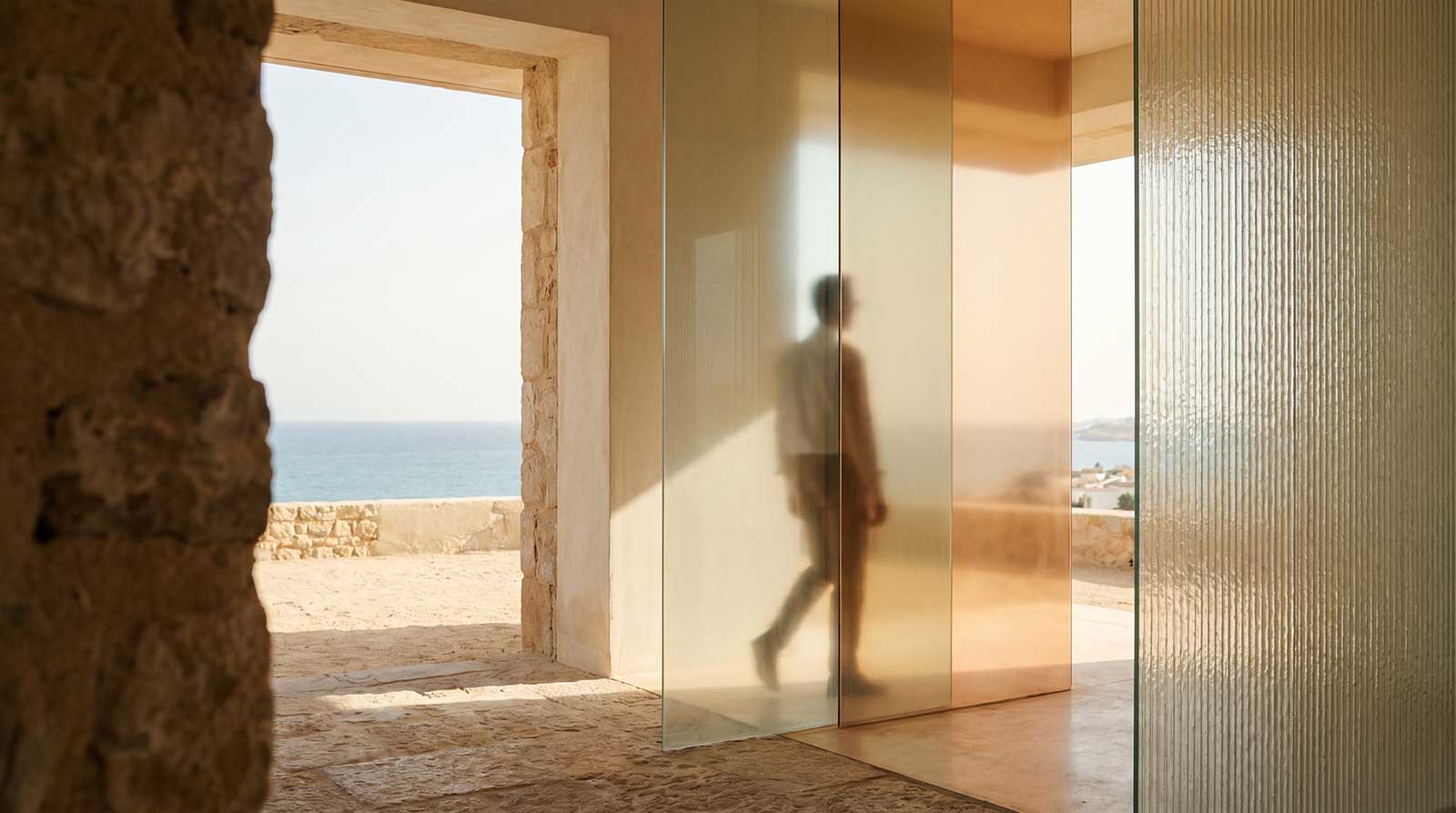 Silueta borrosa de una persona caminando detrás de paneles de vidrio translúcido con vista al mar y paredes de piedra.