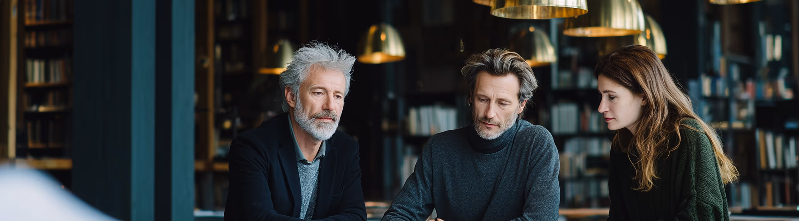 Trois personnes concentrées travaillant ensemble dans une bibliothèque moderne avec des lampes suspendues.
