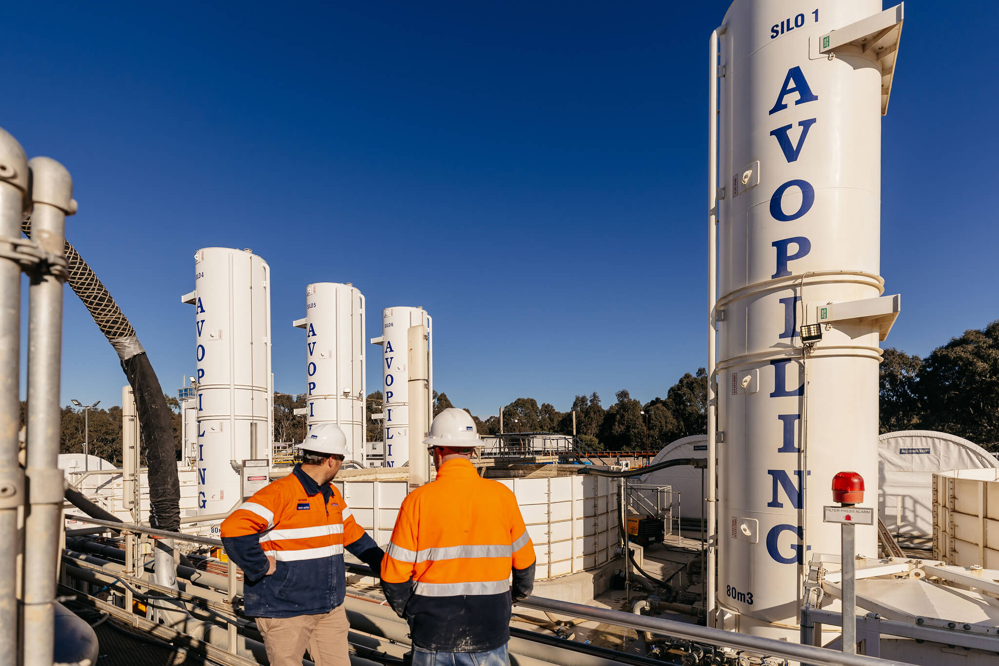 White industrial silos labeled 'AVOPIL' at a construction site with workers and machinery in the background.
