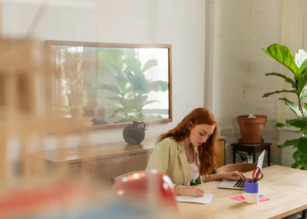 Person writing notes at a desk next to a laptop in a bright office