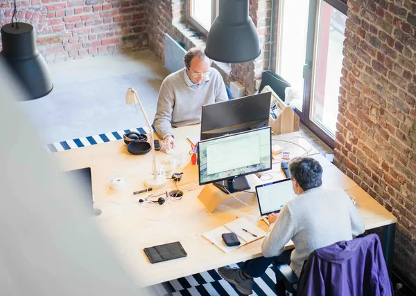 Two people working at computers at a shared desk in a bright office with brick walls