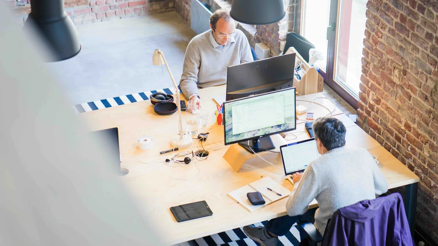 Two people working at computers at a shared desk in a bright office with brick walls