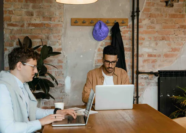 Two people working on laptops at a wooden table in an office with brick walls