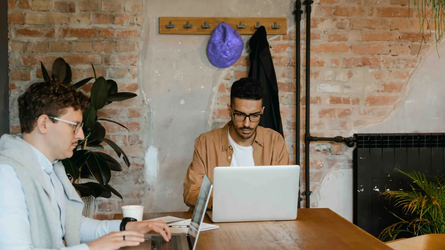 Two people working on laptops at a wooden table in an office with brick walls