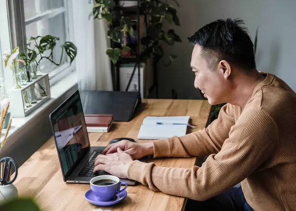 Person typing on a laptop at a wooden desk by a window, with a purple coffee cup in front