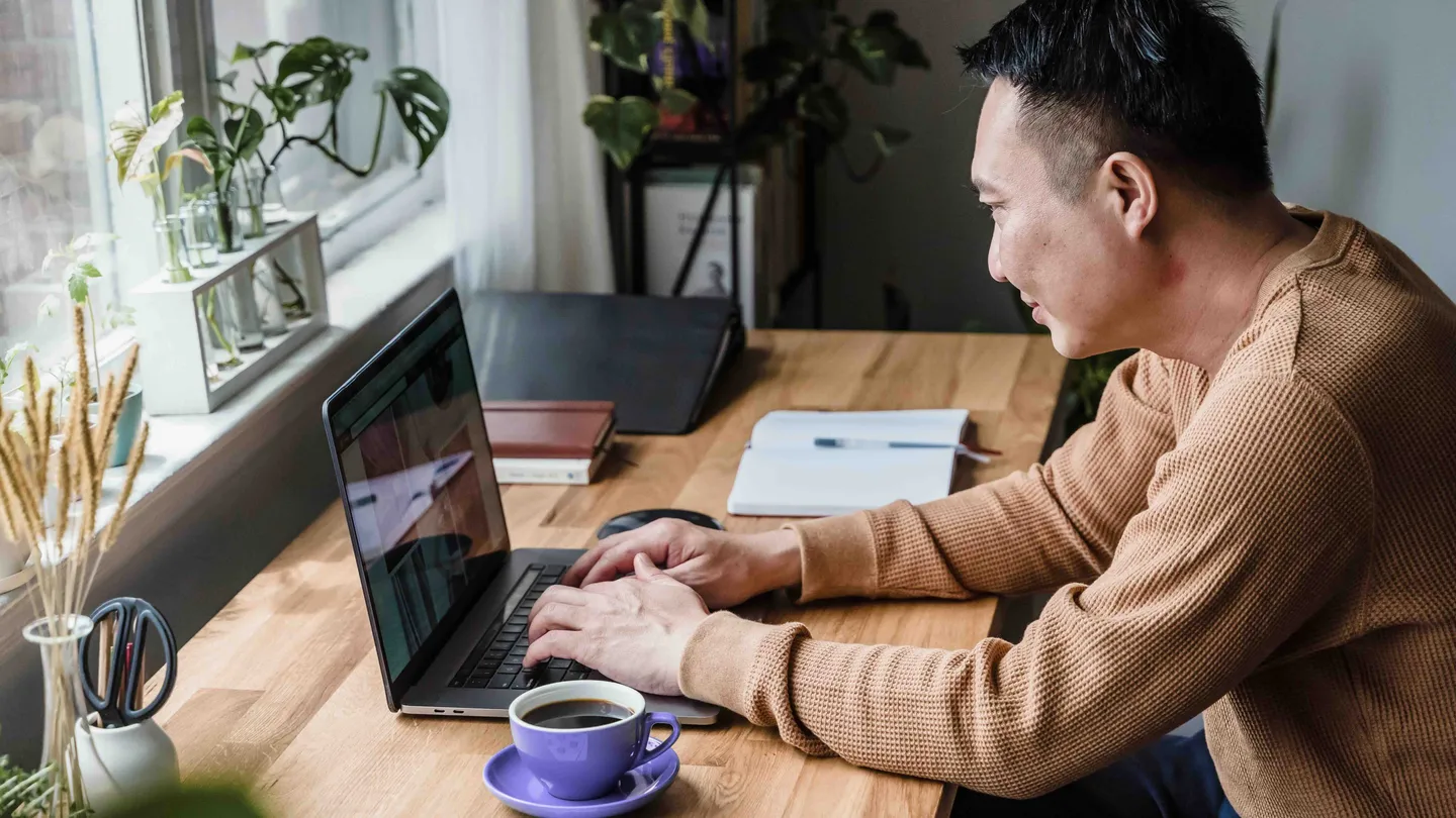 Person typing on a laptop at a wooden desk by a window, with a purple coffee cup in front