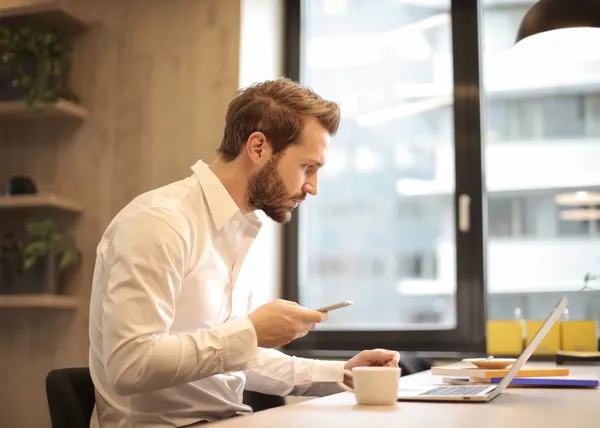 Person at a desk looking at a phone next to an open laptop and a coffee cup by a large window