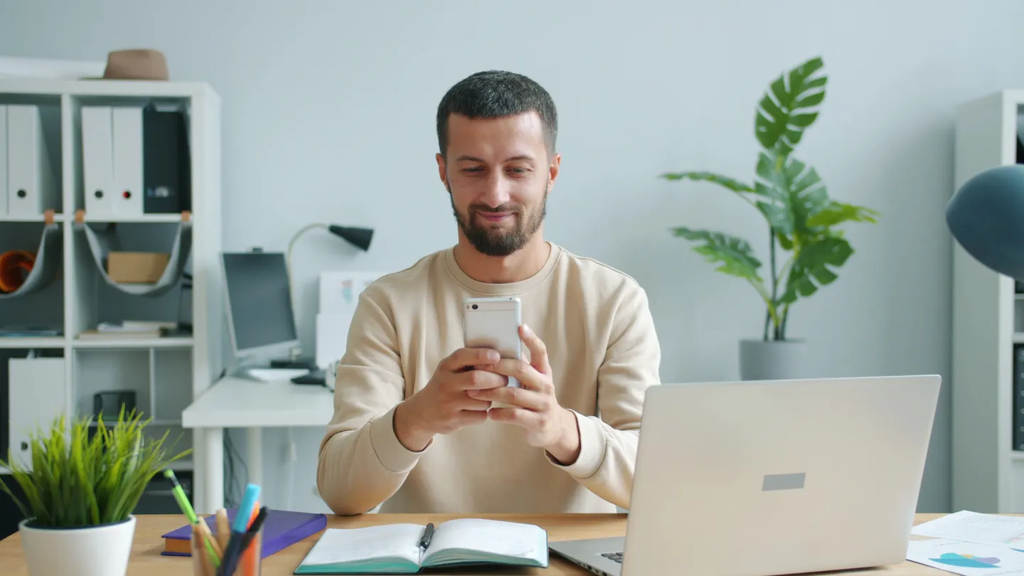 Smiling person looking at a phone at a desk with an open laptop and notebook in a bright office