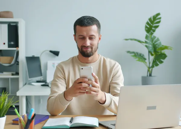 Smiling person looking at a phone at a desk with an open laptop and notebook in a bright office