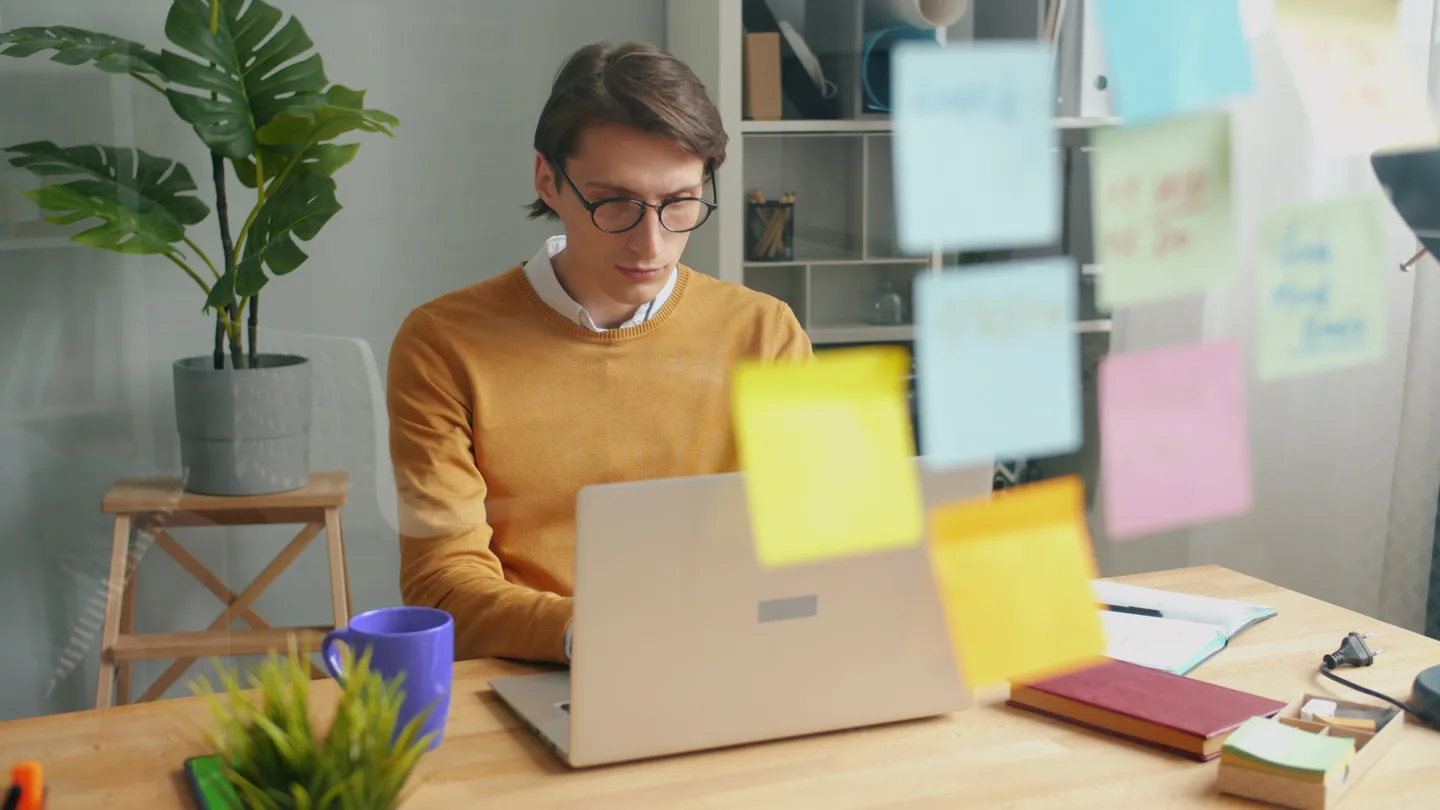 Person working on a laptop at a desk with a purple mug and sticky notes on a glass wall