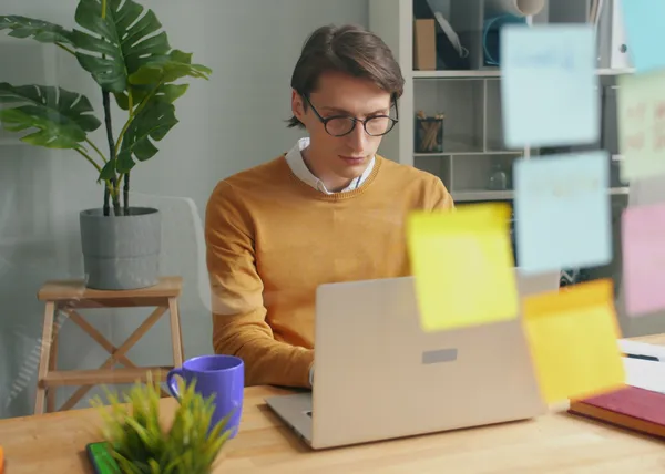 Person working on a laptop at a desk with a purple mug and sticky notes on a glass wall