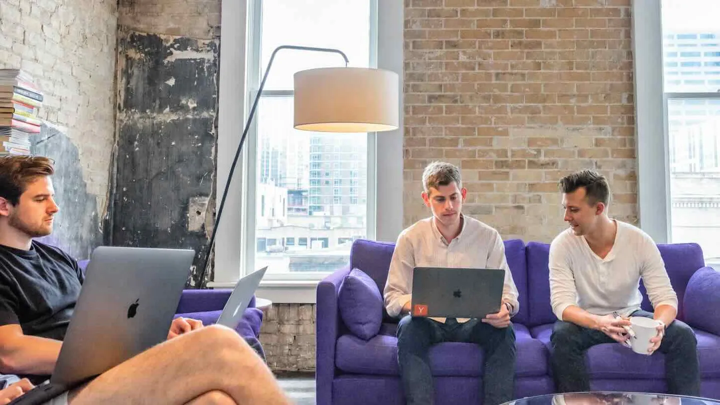 Three people working on laptops on purple couches in a sunlit office with brick walls