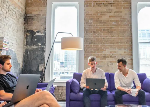Three people working on laptops on purple couches in a sunlit office with brick walls