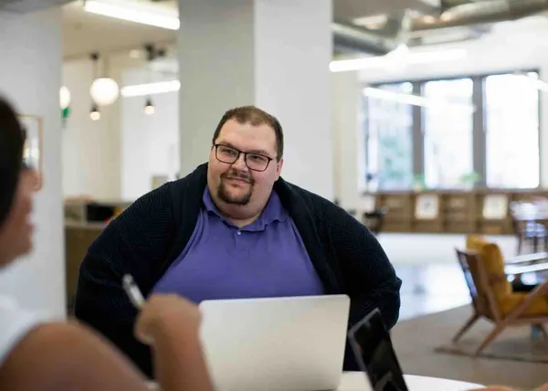 Smiling person in glasses and a navy cardigan sitting at a laptop across a table from a colleague in a bright office