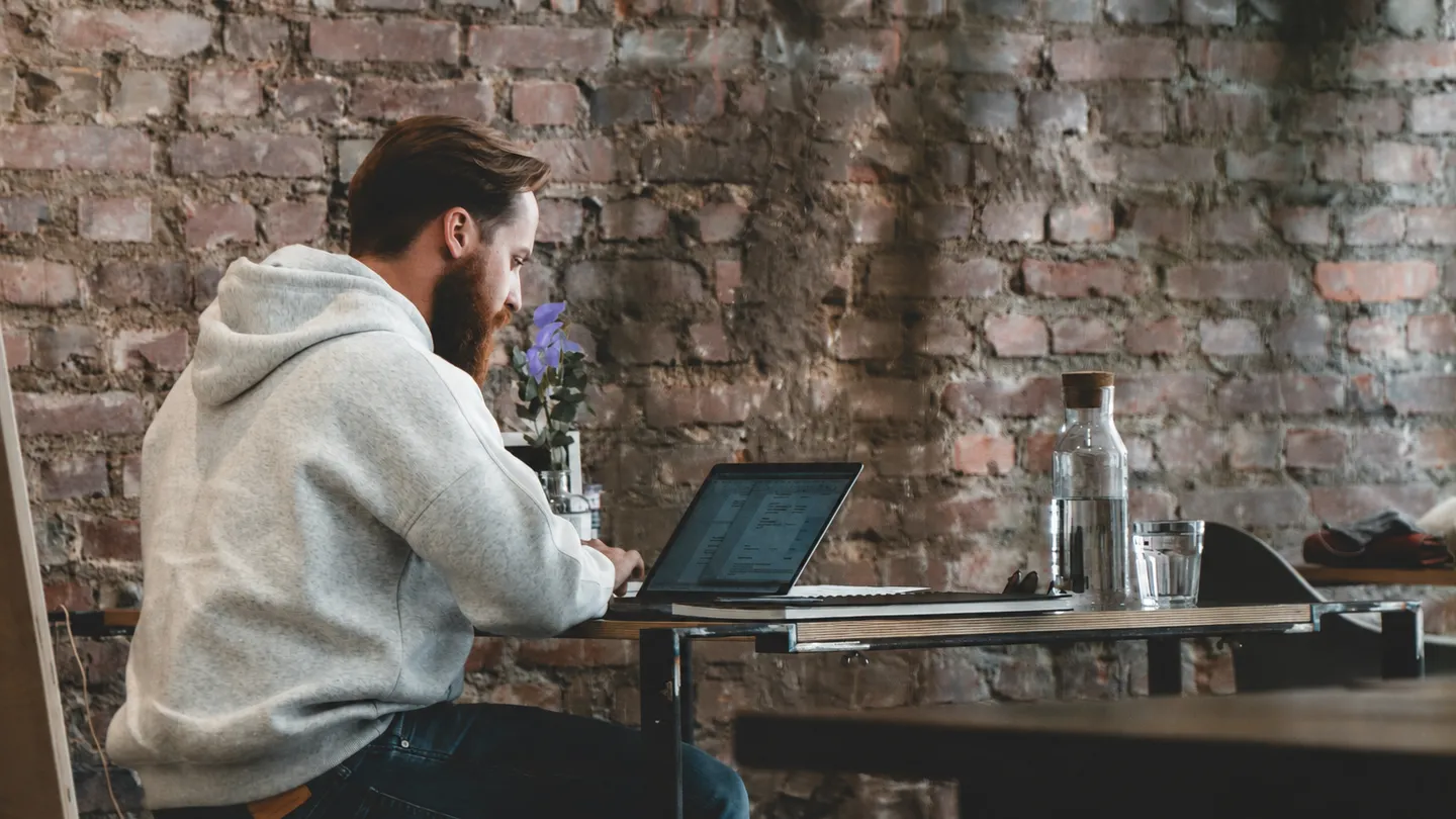 Person with a beard in a gray hoodie working on a laptop at a table against a brick wall