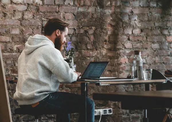 Person with a beard in a gray hoodie working on a laptop at a table against a brick wall