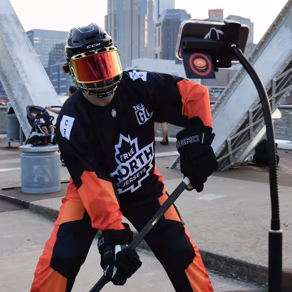 Person in black and orange hockey gear holding a hockey stick, standing on an urban bridge with skyscrapers in the background.