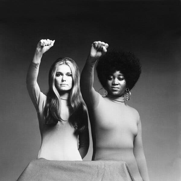 A black-and-white photograph of Gloria Steinem, with long hair, and Dorothy Pitman Hughes, with an Afro, both in light turtlenecks and holding their fists upward.