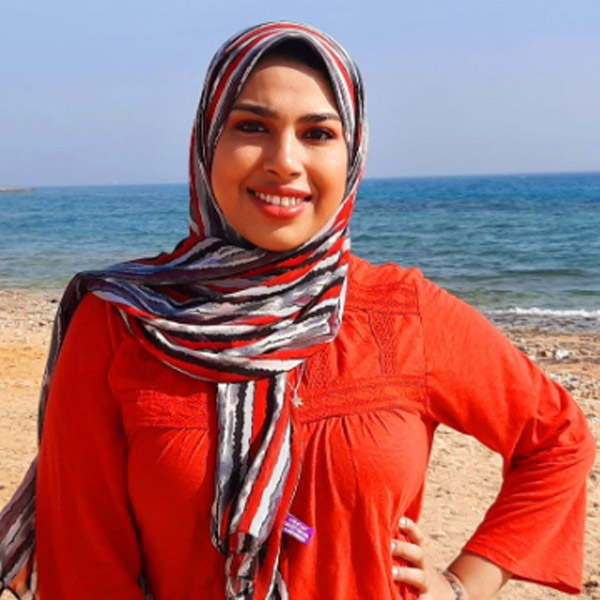 Headshot of Fatma Tawfik smiling, wearing a red top and patterned headscarf near the ocean.