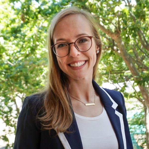 Headshot of Katie Usalis smiling outdoors, wearing glasses and a navy blazer.