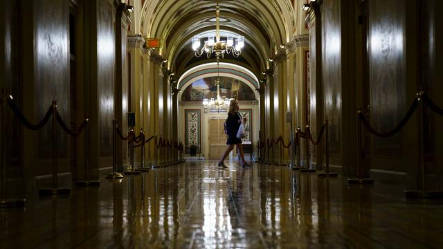 A lone woman walks down a dimly lit, ornate arched corridor of the US Capitol, her figure silhouetted against warm light reflecting off the polished floor.