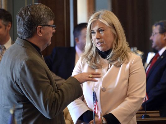 A man in a brown jacket gestures while speaking with a blonde woman in a pale pink blazer inside a legislative chamber, small American flags visible nearby.