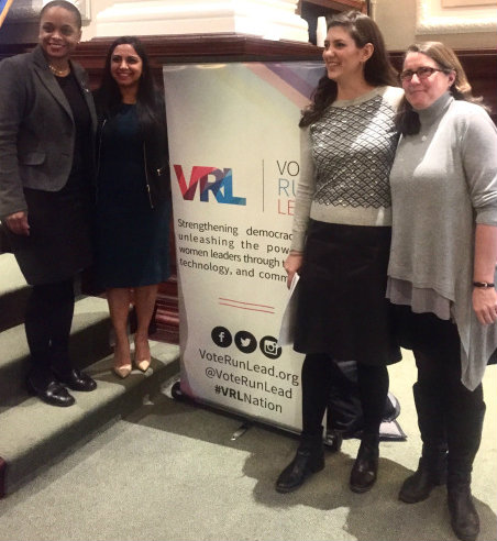 Four women in professional attire smile and pose together beside a tall Vote Run Lead banner promoting women's leadership, in an ornate stair-lined hall.