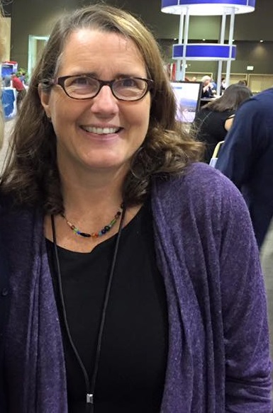 Cynthia Terrell: A smiling woman with glasses, wavy brown hair, and a purple cardigan over a black top stands at a busy indoor conference venue with booths blurred behind her.