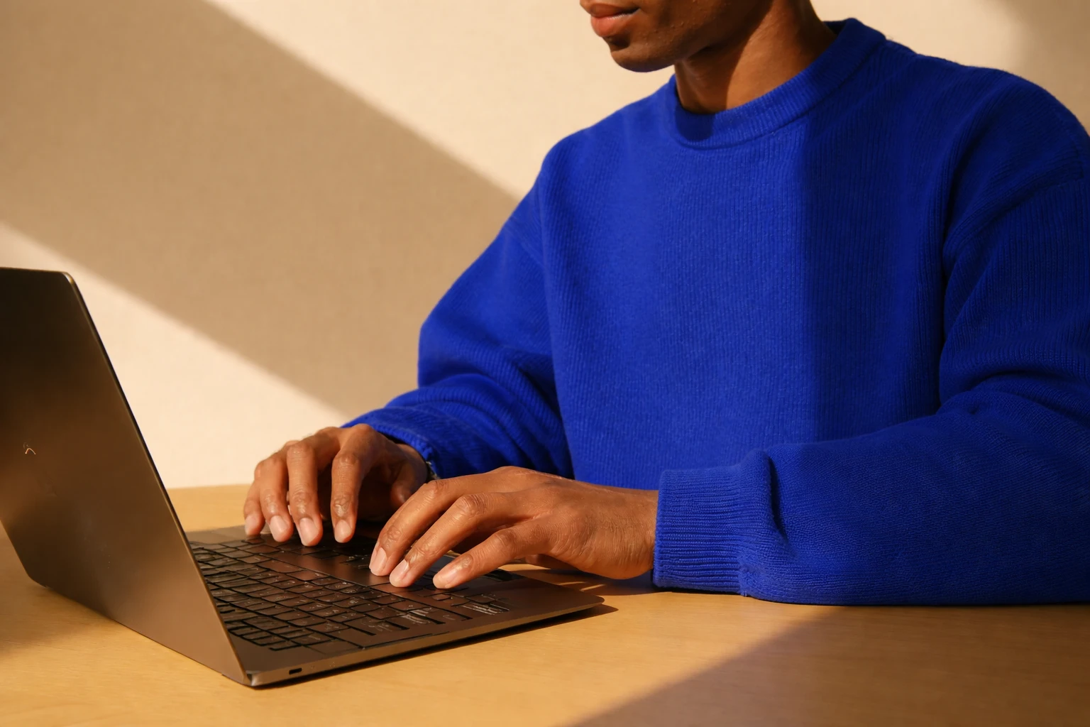 Person wearing a blue sweater typing on a laptop at a wooden table with sunlight casting shadows.