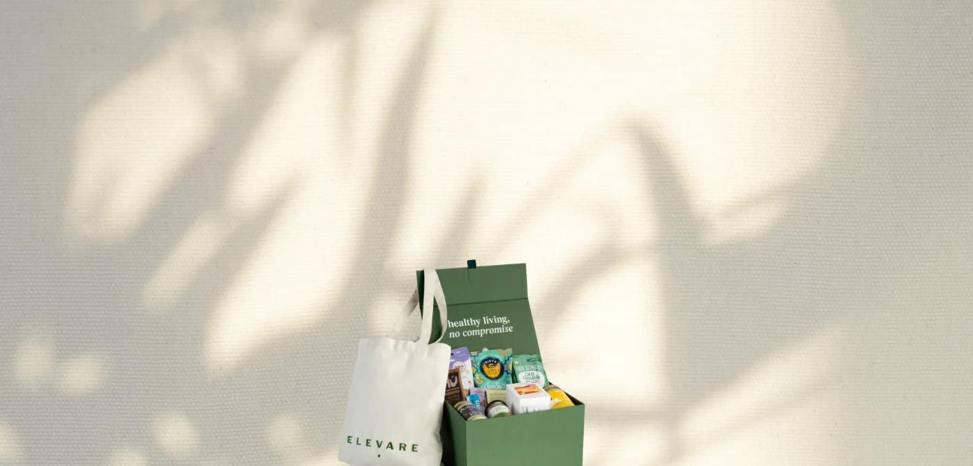 Green box with healthy food and wellness products and a white tote bag with ELEVARE text, set against a textured wall with natural shadows.