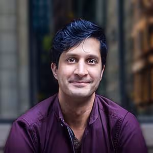 Man with short dark hair wearing a maroon shirt, smiling softly with a blurred background.