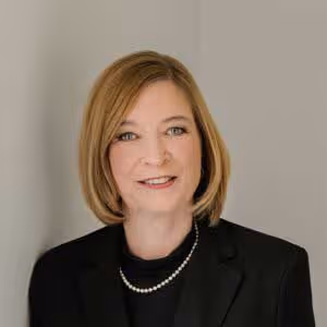 Professional headshot of a woman with shoulder-length blonde hair wearing a black blazer and pearl necklace against a neutral background.