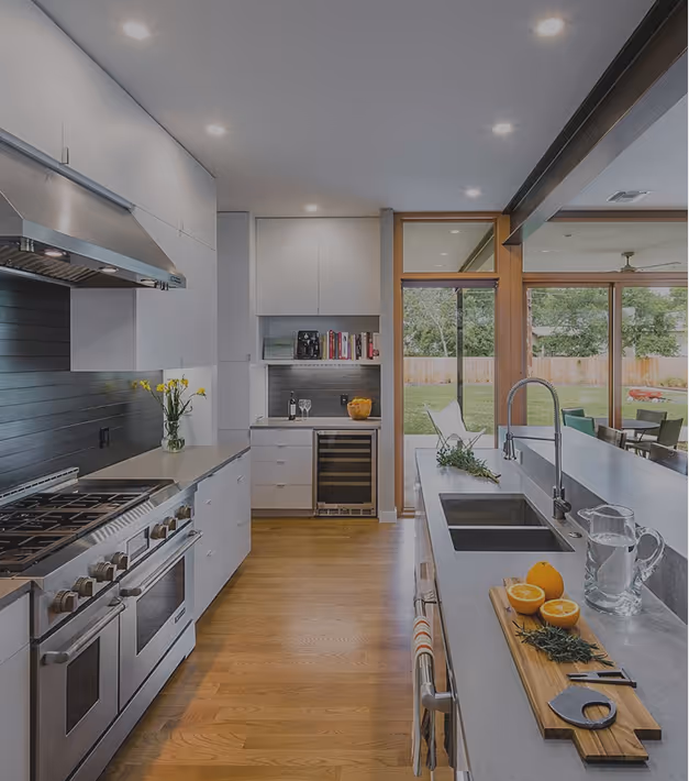 Modern kitchen with stainless steel stove, gray countertops, wooden floor, and large windows overlooking a green backyard.