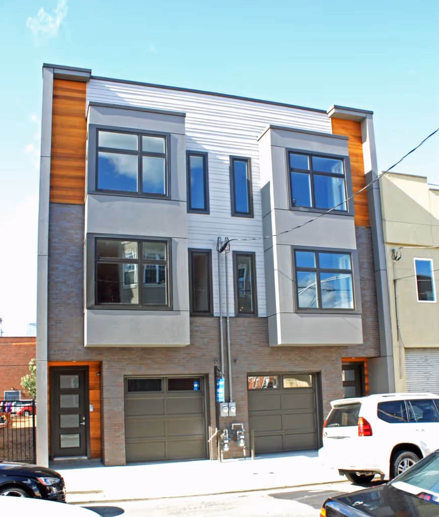 Modern two-story duplex with gray garage doors, large windows, and wood accents, situated in an urban neighborhood.