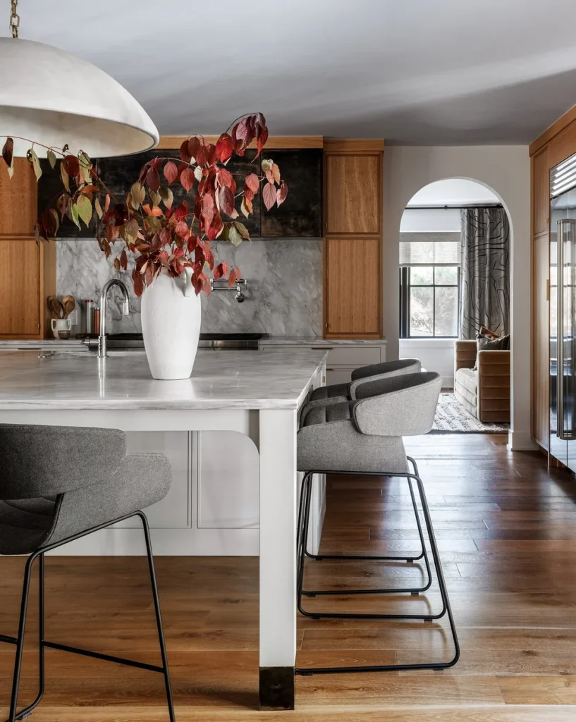 Modern kitchen with white marble island, gray upholstered bar stools, wooden cabinets, and a white vase with red and green leaves centerpiece.