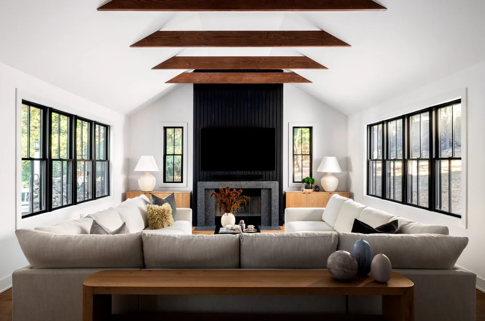 Modern living room with white sofas, wooden beams on ceiling, large windows, and a black fireplace with a mounted TV.