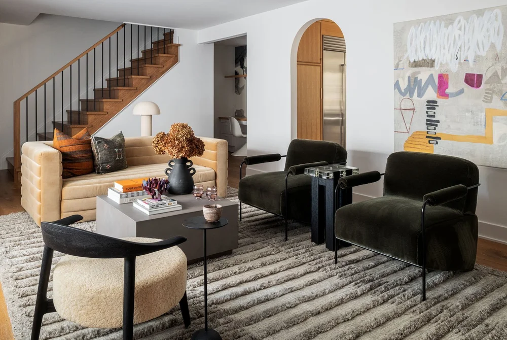 Modern living room with beige sofa, two dark green armchairs, a black and cream accent chair, and a gray textured rug under a wooden staircase.