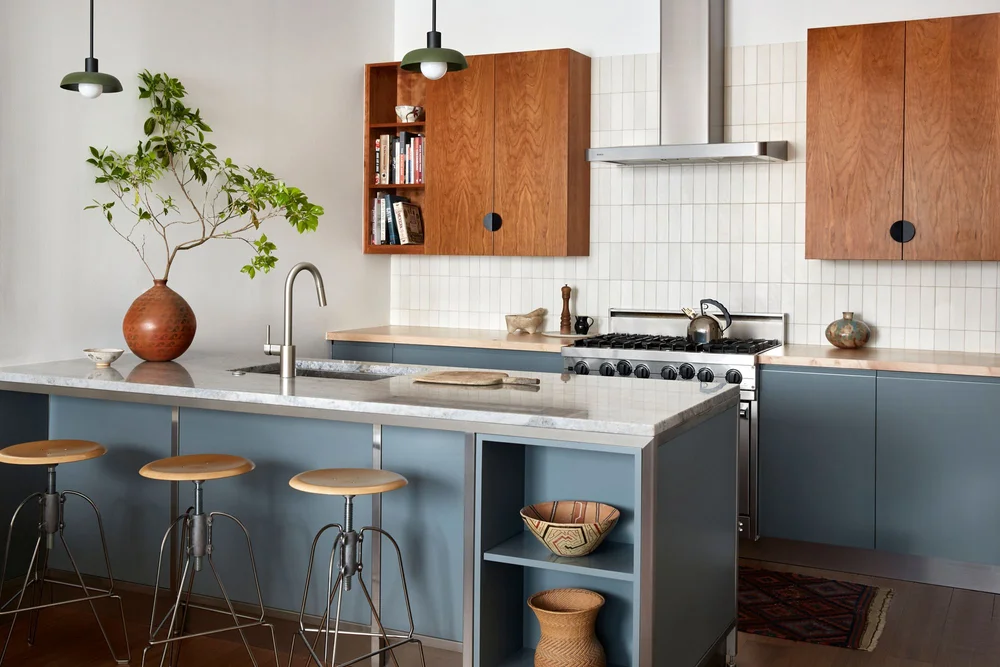 Modern kitchen with blue island counter, marble countertop, wooden upper cabinets, stainless steel stove, and a large pottery vase with green leafy branches.