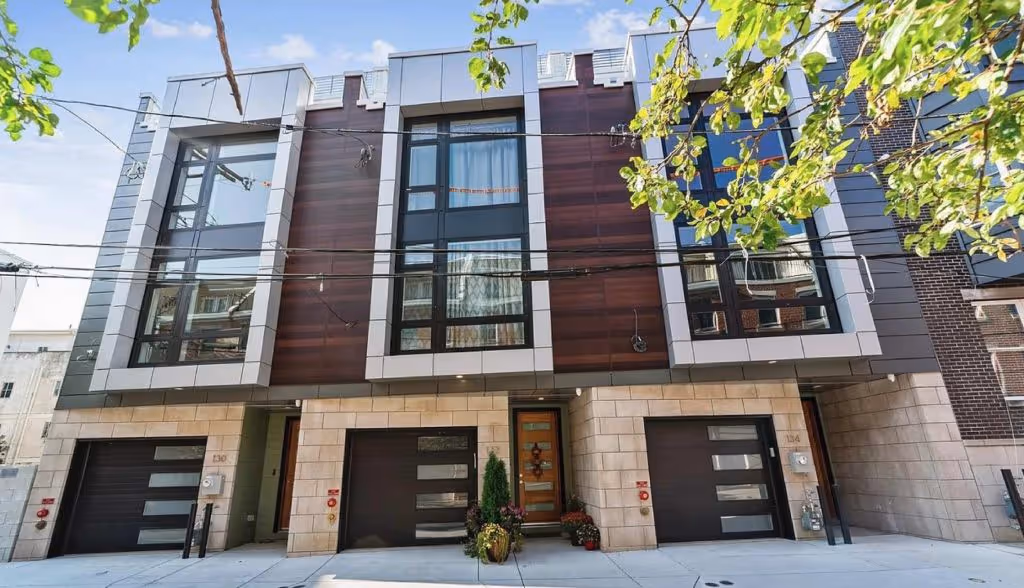 Modern three-story townhouse with large windows, three garage doors, and a wooden front door framed by plants.