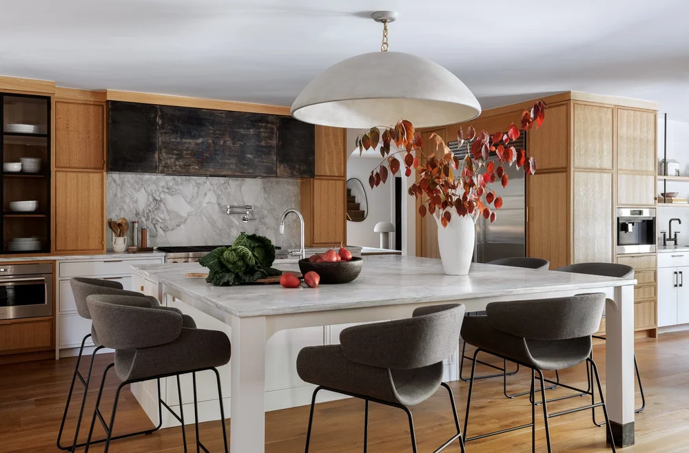 Modern kitchen with marble island, six gray cushioned stools, wooden cabinets, stainless steel appliances, and a large white pendant light above decorated with autumn leaves in a white vase.
